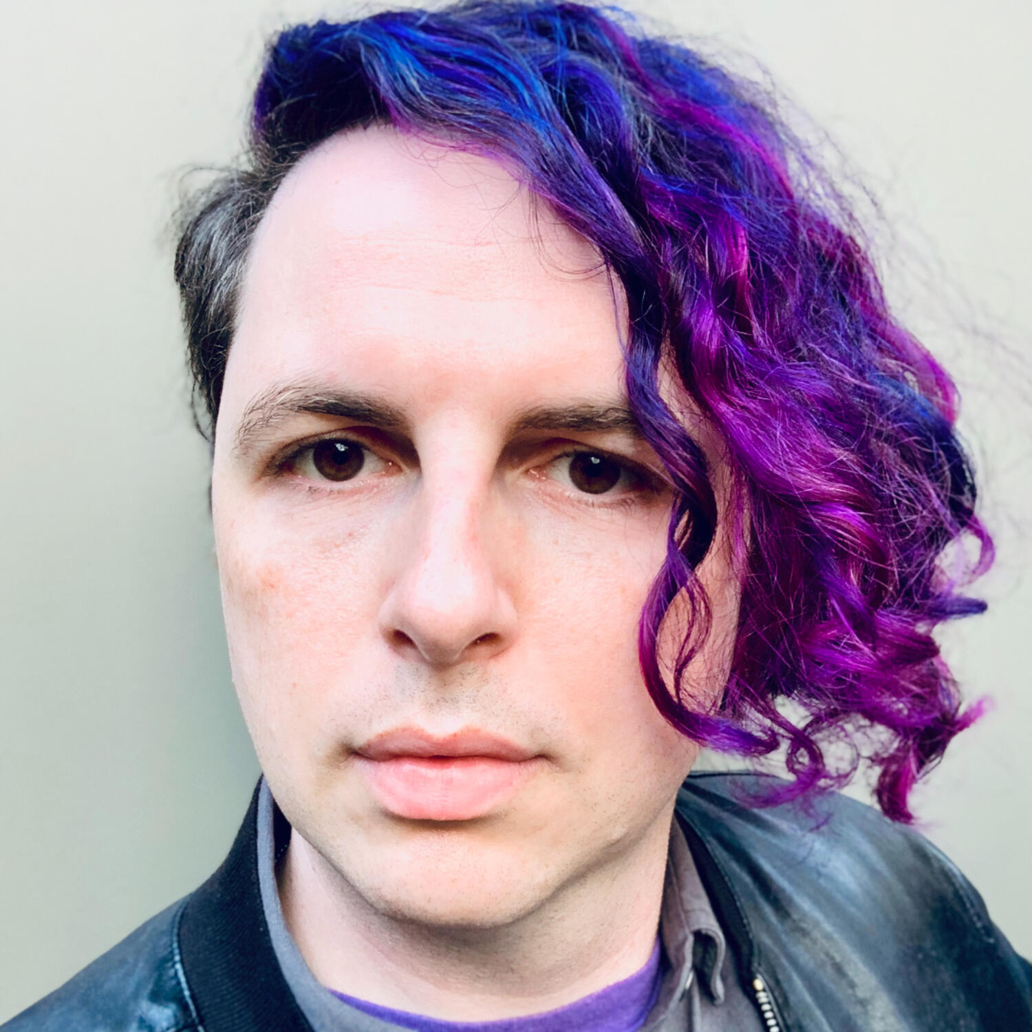 Tom Lowenthal — a pale white person with brown eyes rosy cheeks looks directly at you. Their asymmetric hair is purple with highlights of blue and magenta.